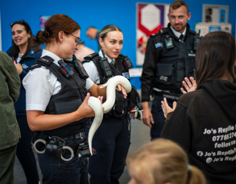 Police officers in uniform speak with members of the public at an indoor event, as one officer holds a white snake and shows it to a small group. A person wearing a black hoodie with “Jo’s Reptile Parties” on the back stands nearby, while other attendees are visible in the background.