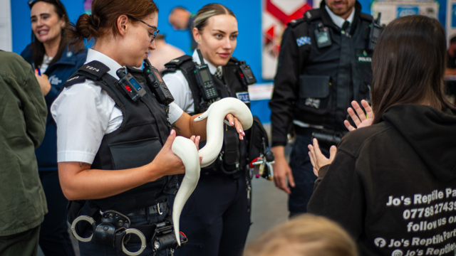 Police officers in uniform speak with members of the public at an indoor event, as one officer holds a white snake and shows it to a small group. A person wearing a black hoodie with “Jo’s Reptile Parties” on the back stands nearby, while other attendees are visible in the background.