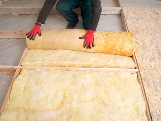 Person wearing red gloves and green pants kneeling on a wooden surface, unrolling yellow fiberglass insulation between wooden beams. Person wearing red gloves and green pants kneeling on a wooden surface, unrolling yellow fiberglass insulation between wooden beams.