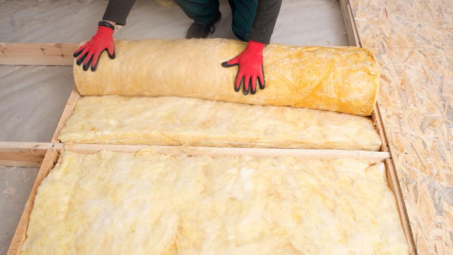 Person wearing red gloves and green pants kneeling on a wooden surface, unrolling yellow fiberglass insulation between wooden beams. Person wearing red gloves and green pants kneeling on a wooden surface, unrolling yellow fiberglass insulation between wooden beams.