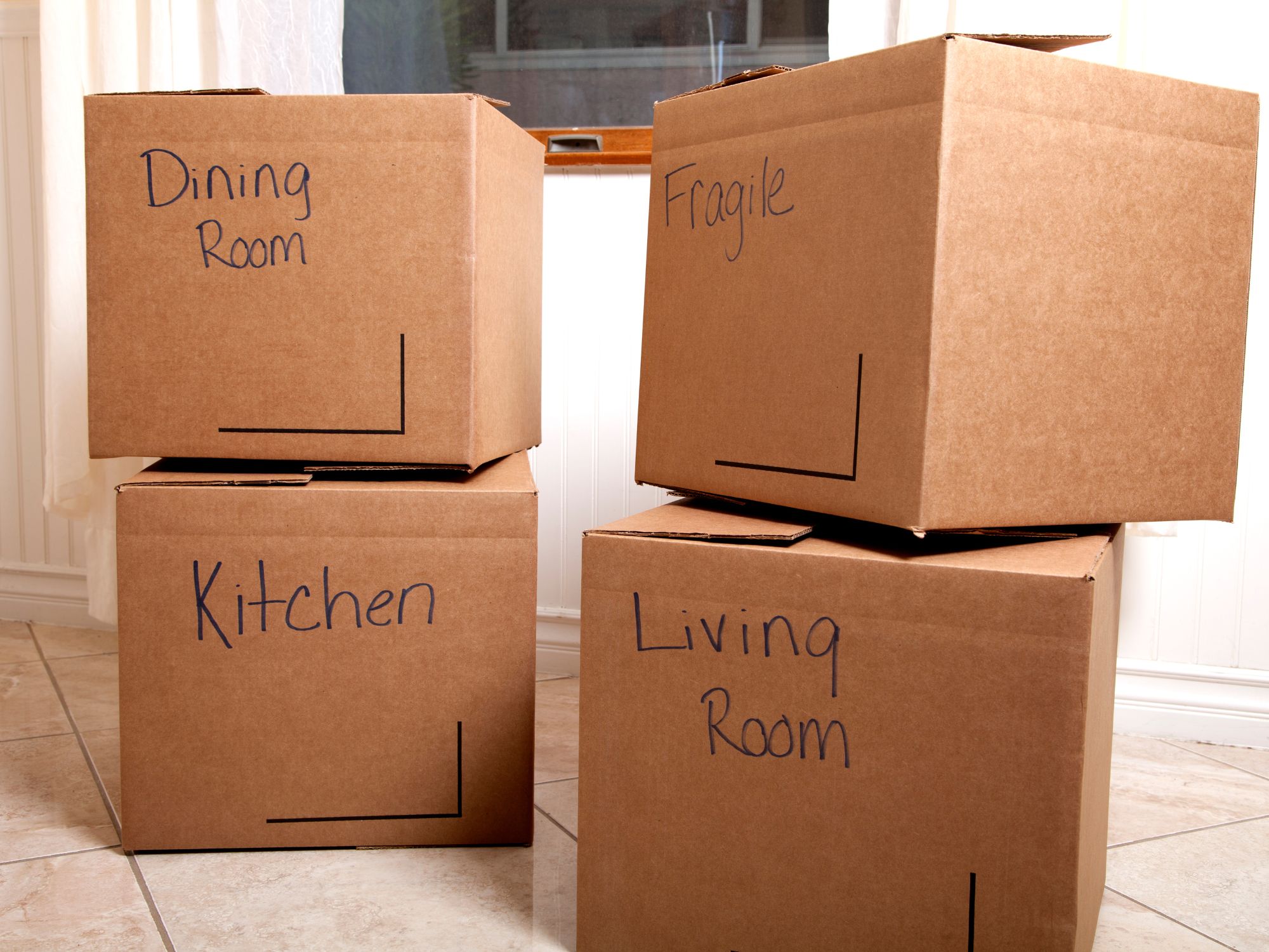 Four labeled cardboard boxes stacked in a room, with handwritten markings reading 'Dining Room', 'Fragile', 'Kitchen', and 'Living Room'—representing a household move or packing process.