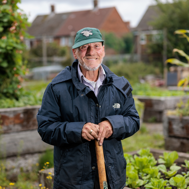 Person in a green cap and blue jacket holding a gardening tool, standing in a garden with raised beds and houses in the background.