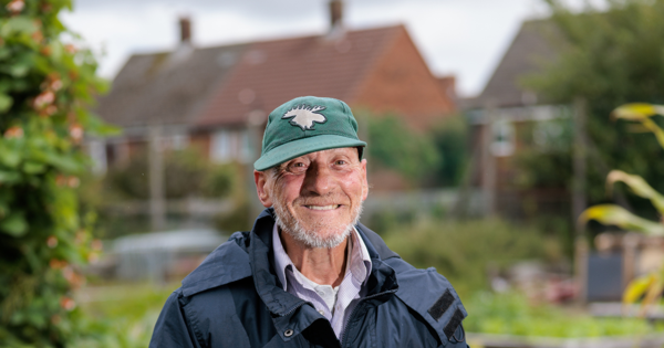 Person in a green cap and blue jacket holding a gardening tool, standing in a garden with raised beds and houses in the background.