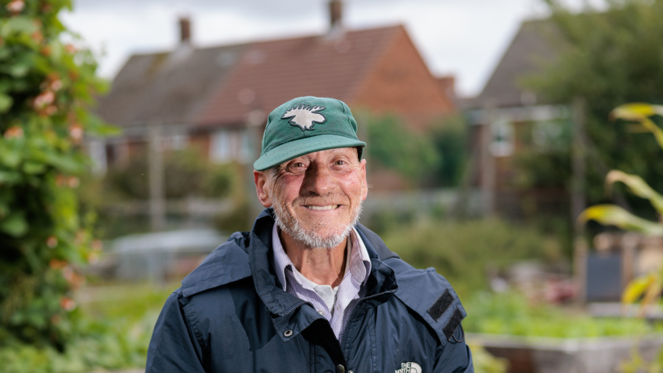Person in a green cap and blue jacket holding a gardening tool, standing in a garden with raised beds and houses in the background. Person in a green cap and blue jacket holding a gardening tool, standing in a garden with raised beds and houses in the background.