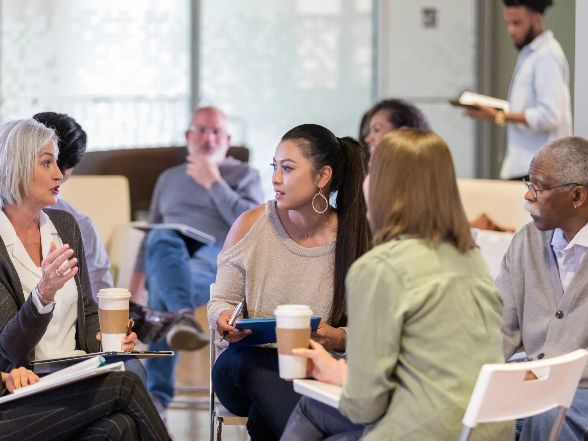 A group of people seated in a circle during an informal workshop, actively engaged in discussion with notebooks, tablets, and coffee cups, reflecting a collaborative and interactive environment.
