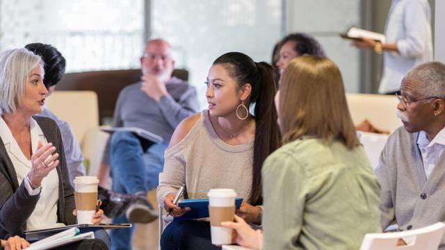 A group of people seated in a circle during an informal workshop, actively engaged in discussion with notebooks, tablets, and coffee cups, reflecting a collaborative and interactive environment.