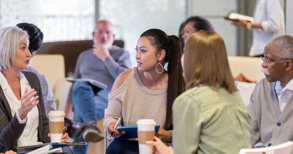 A group of people seated in a circle during an informal workshop, actively engaged in discussion with notebooks, tablets, and coffee cups, reflecting a collaborative and interactive environment.