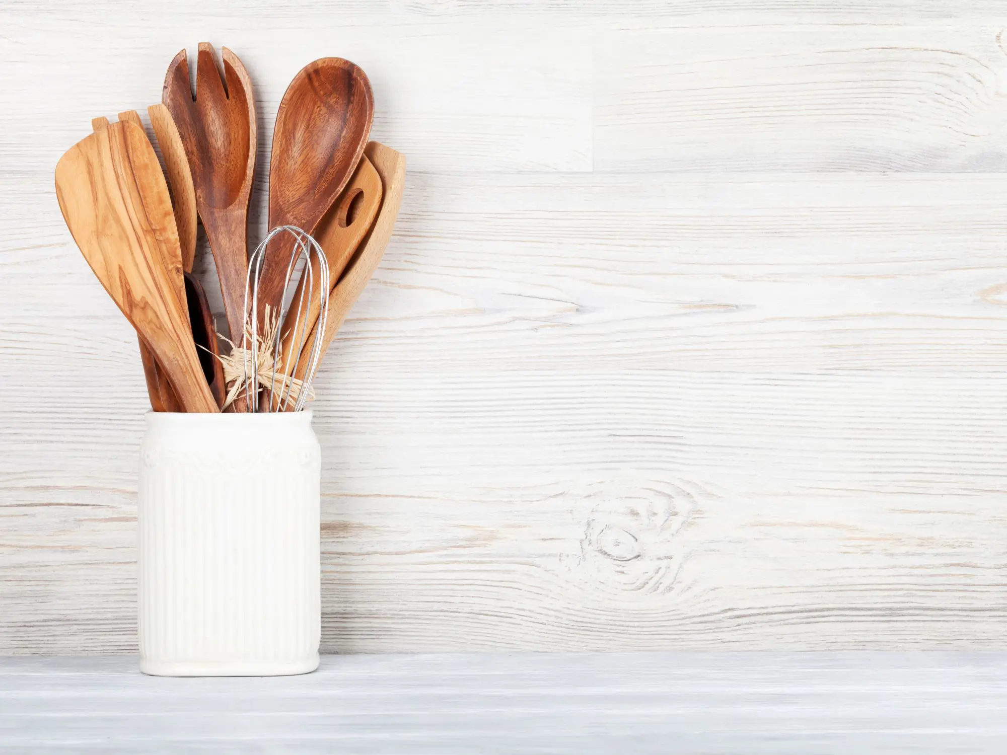 White ceramic container holding wooden kitchen utensils and a metal whisk on a light wooden surface.