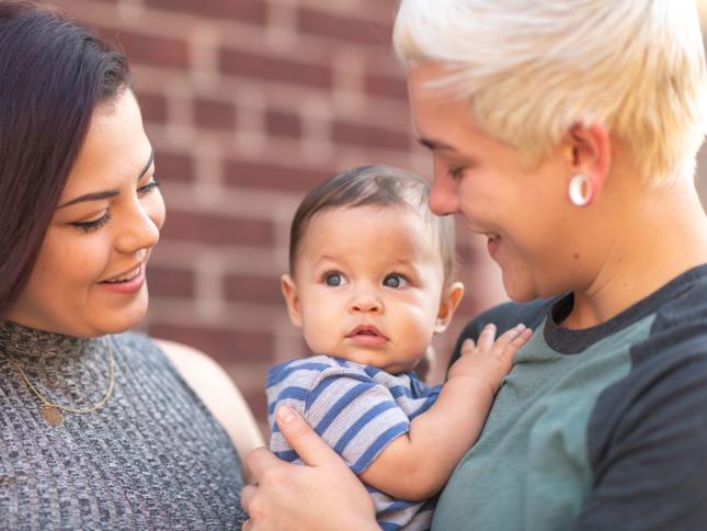 Two adults holding a baby between them in front of a brick wall, representing a young family preparing for homeownership through rent to buy. Two adults holding a baby between them in front of a brick wall, representing a young family preparing for homeownership through rent to buy.