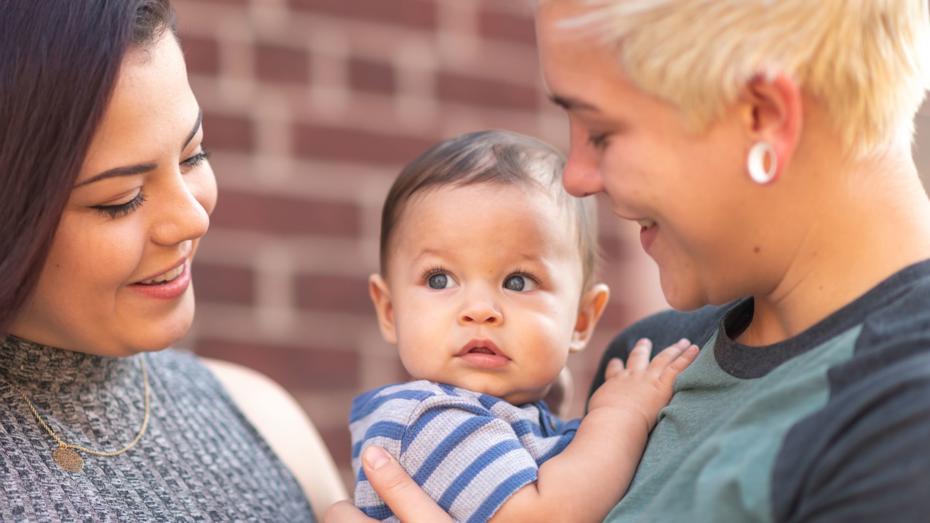 Two adults holding a baby between them in front of a brick wall, representing a young family preparing for homeownership through rent to buy. Two adults holding a baby between them in front of a brick wall, representing a young family preparing for homeownership through rent to buy.