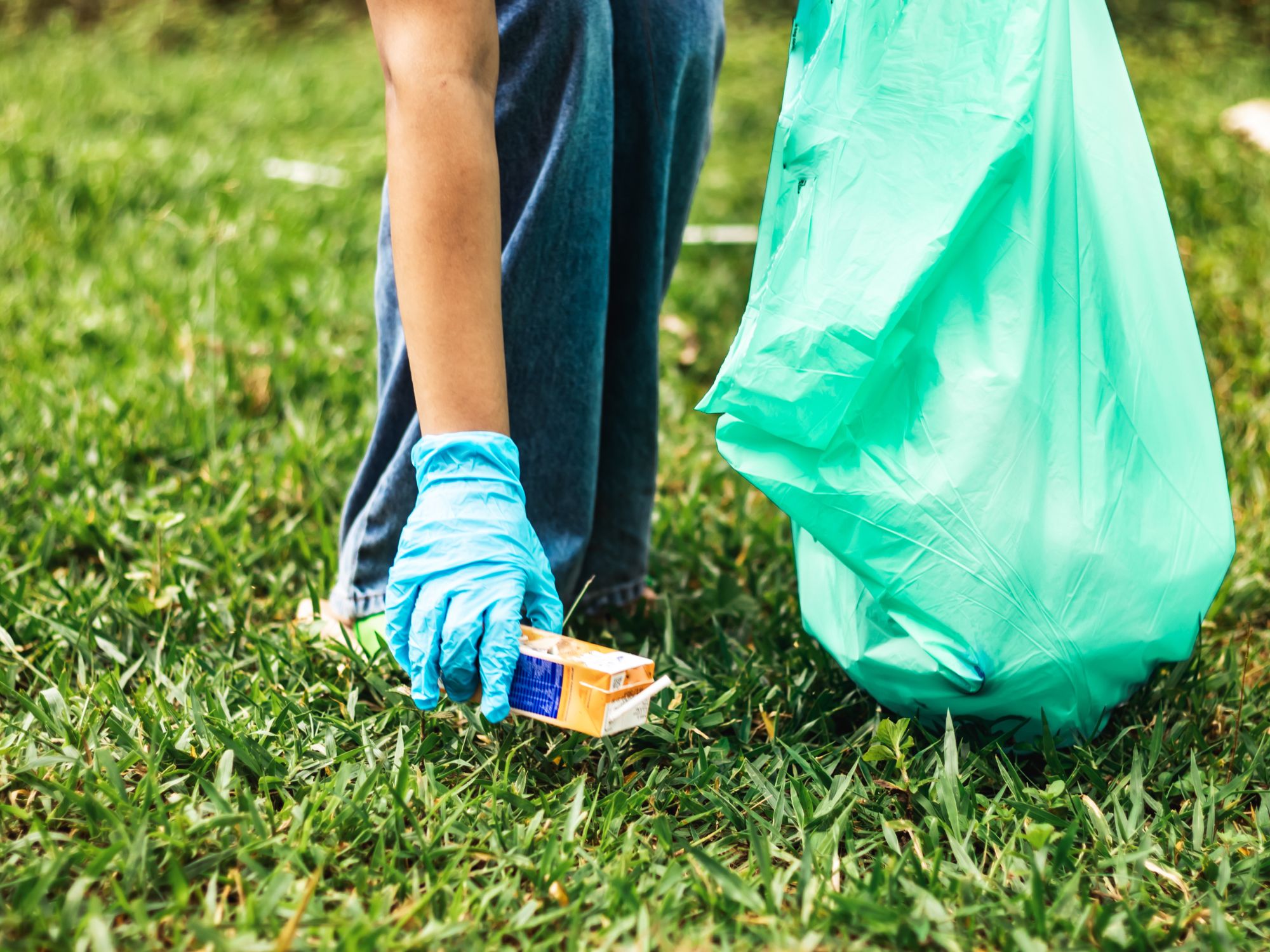 Person wearing blue gloves picking up a small cardboard box from the grass while holding a large green trash bag, suggesting a community or environmental cleanup activity.