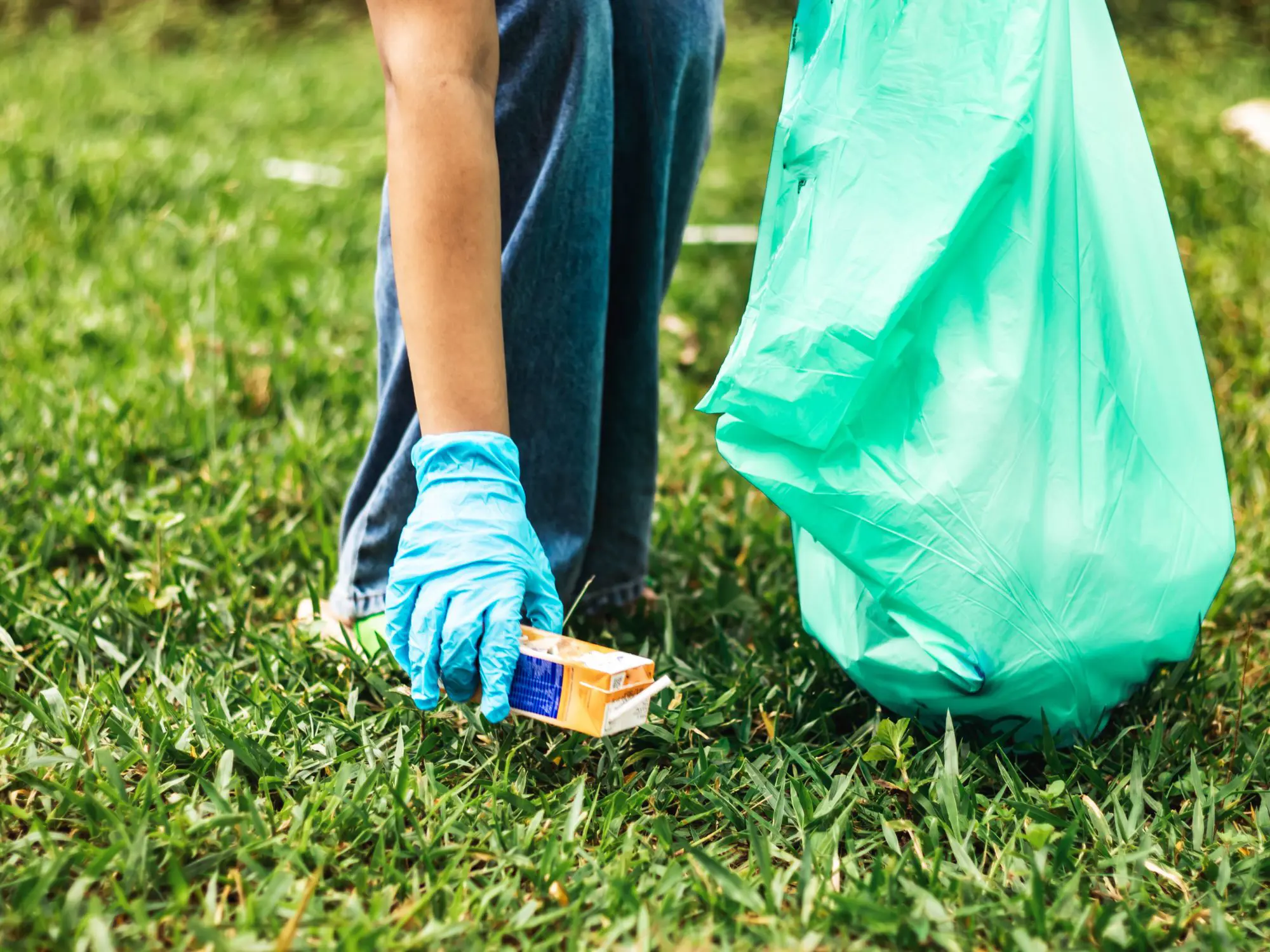 Person wearing blue gloves picking up a small cardboard box from the grass while holding a large green trash bag, suggesting a community or environmental cleanup activity.