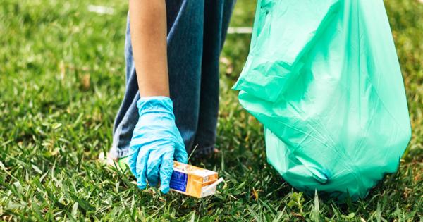 Person wearing blue gloves picking up a small cardboard box from the grass while holding a large green trash bag, suggesting a community or environmental cleanup activity.