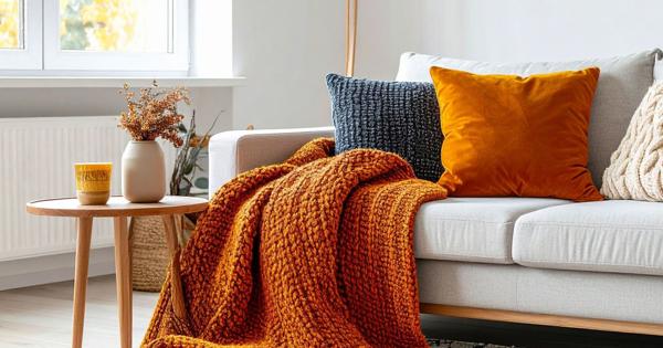 A cozy living room with a light gray sofa, colorful pillows, and an orange knitted throw blanket. A wooden side table holds a yellow mug, dried flowers, and decor, with sunlight streaming through large windows