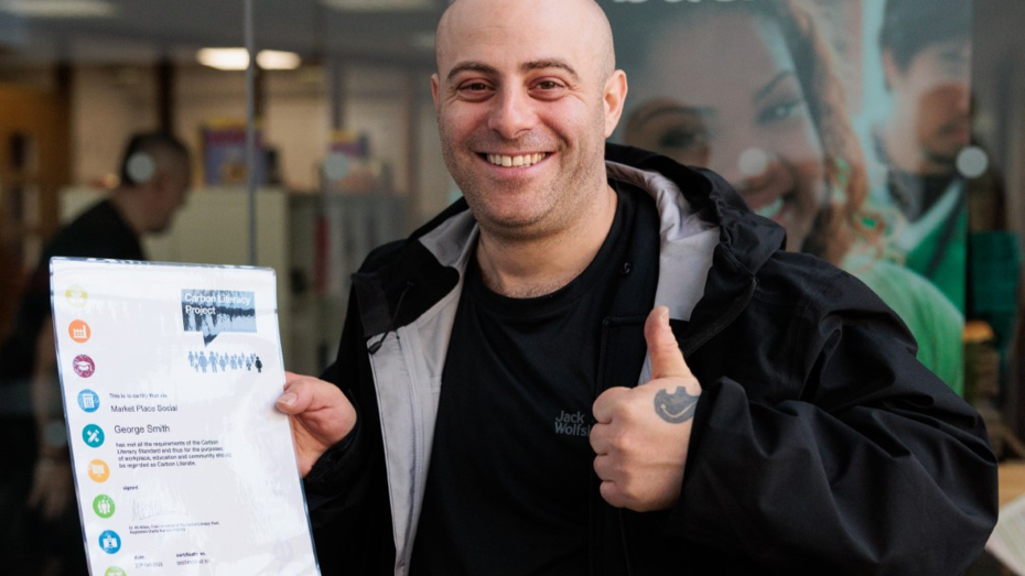 Person standing outdoors holding a certificate and giving a thumbs-up, with glass behind them displaying the words 'Give a little bit back'.