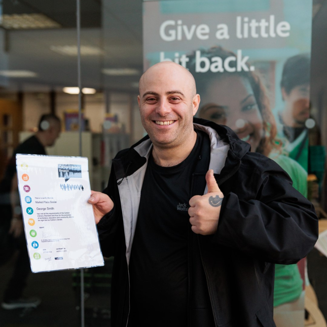 Person standing outdoors holding a certificate and giving a thumbs-up, with glass behind them displaying the words 'Give a little bit back'.