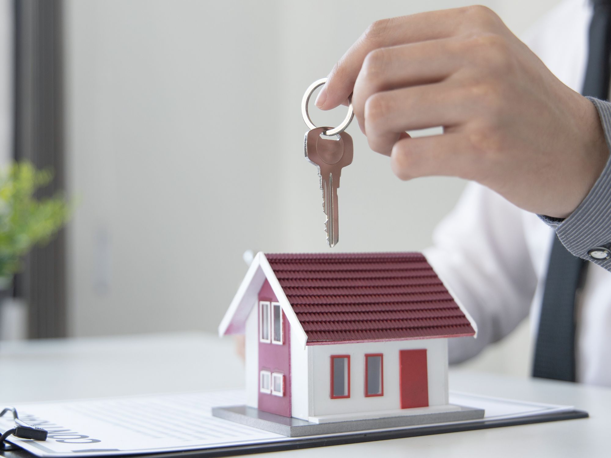 Hand holding a key above a small model house with a red roof, placed on top of property documents.