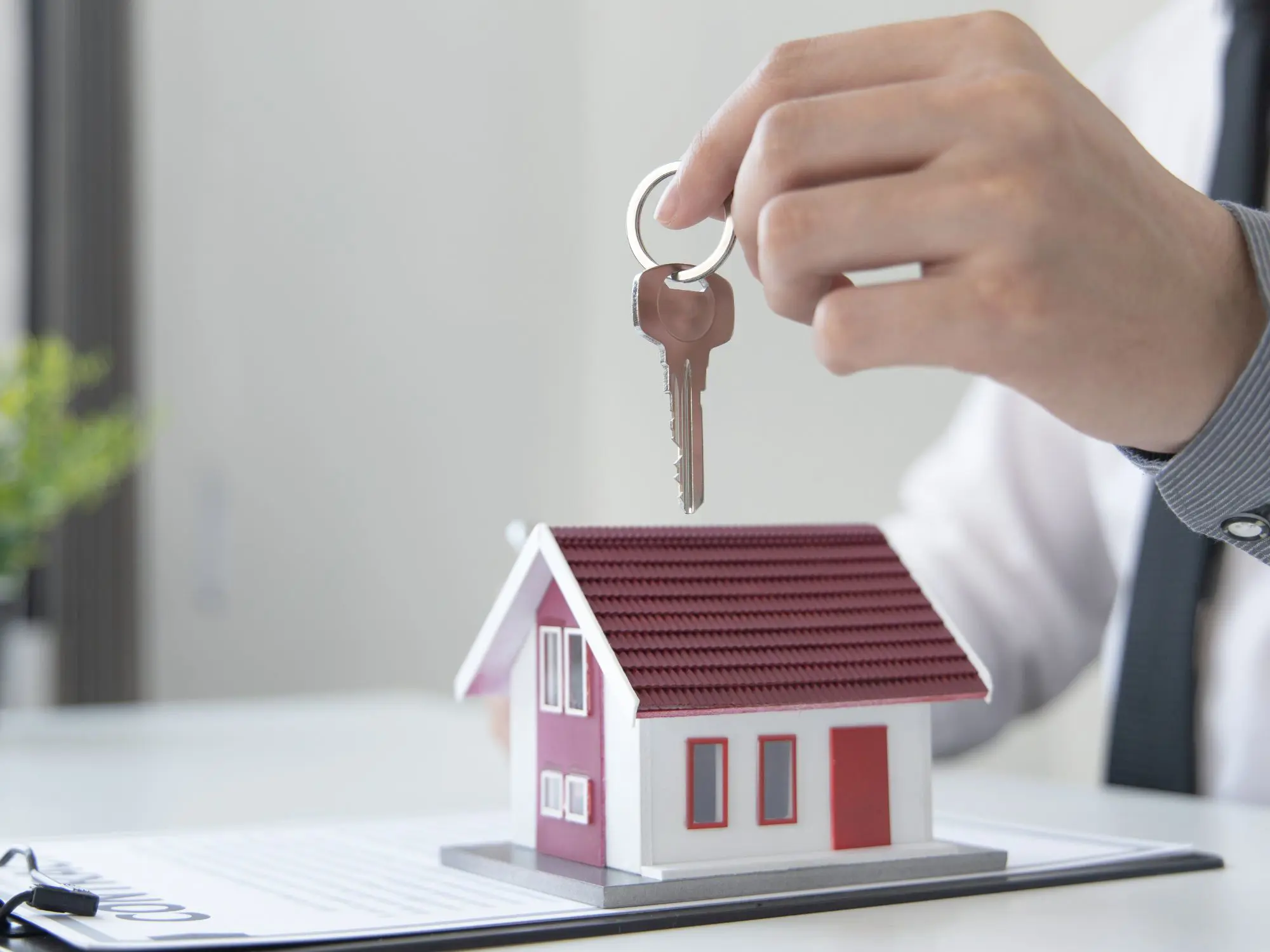 Hand holding a key above a small model house with a red roof, placed on top of property documents.