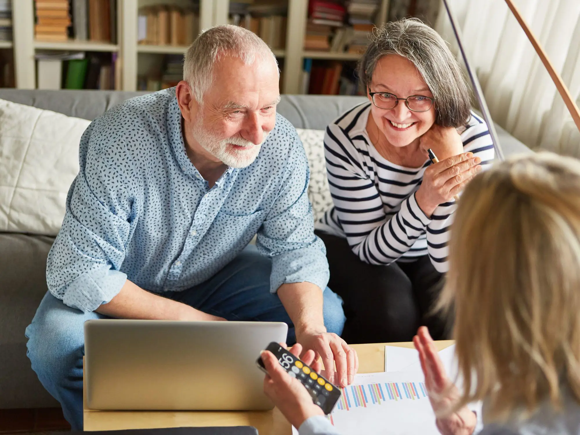 Three people in a room; two sit on a couch, one holds a smartphone with the calculator app open, and a laptop rests on the table in front of them. Bookshelves filled with books line the background.