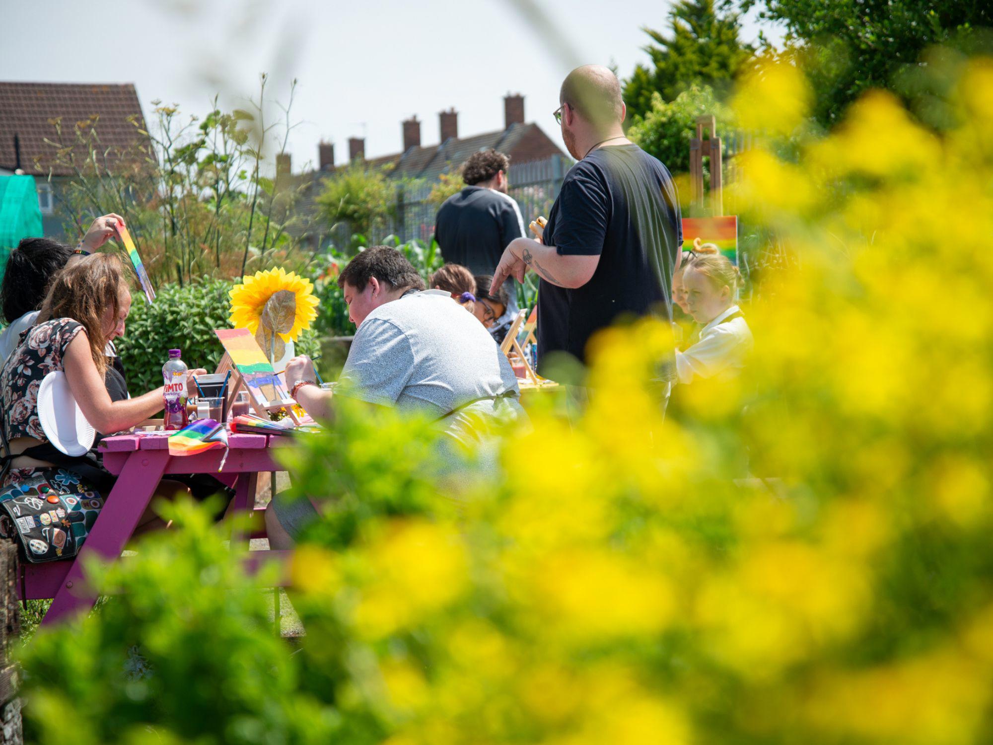 A group of people sitting at a picnic table in a garden, participating in an outdoor art or craft activity with paintbrushes and colourful materials, surrounded by yellow flowers and plants.
