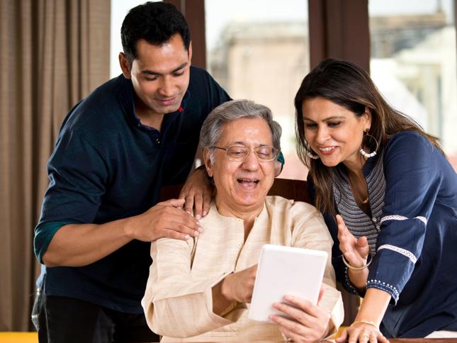 An older person sitting at a table holding a tablet, with two adults standing close by and looking at the screen together in a bright indoor setting. An older person sitting at a table holding a tablet, with two adults standing close by and looking at the screen together in a bright indoor setting.