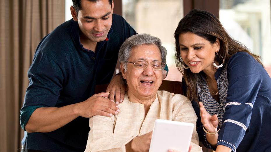 An older person sitting at a table holding a tablet, with two adults standing close by and looking at the screen together in a bright indoor setting. An older person sitting at a table holding a tablet, with two adults standing close by and looking at the screen together in a bright indoor setting.