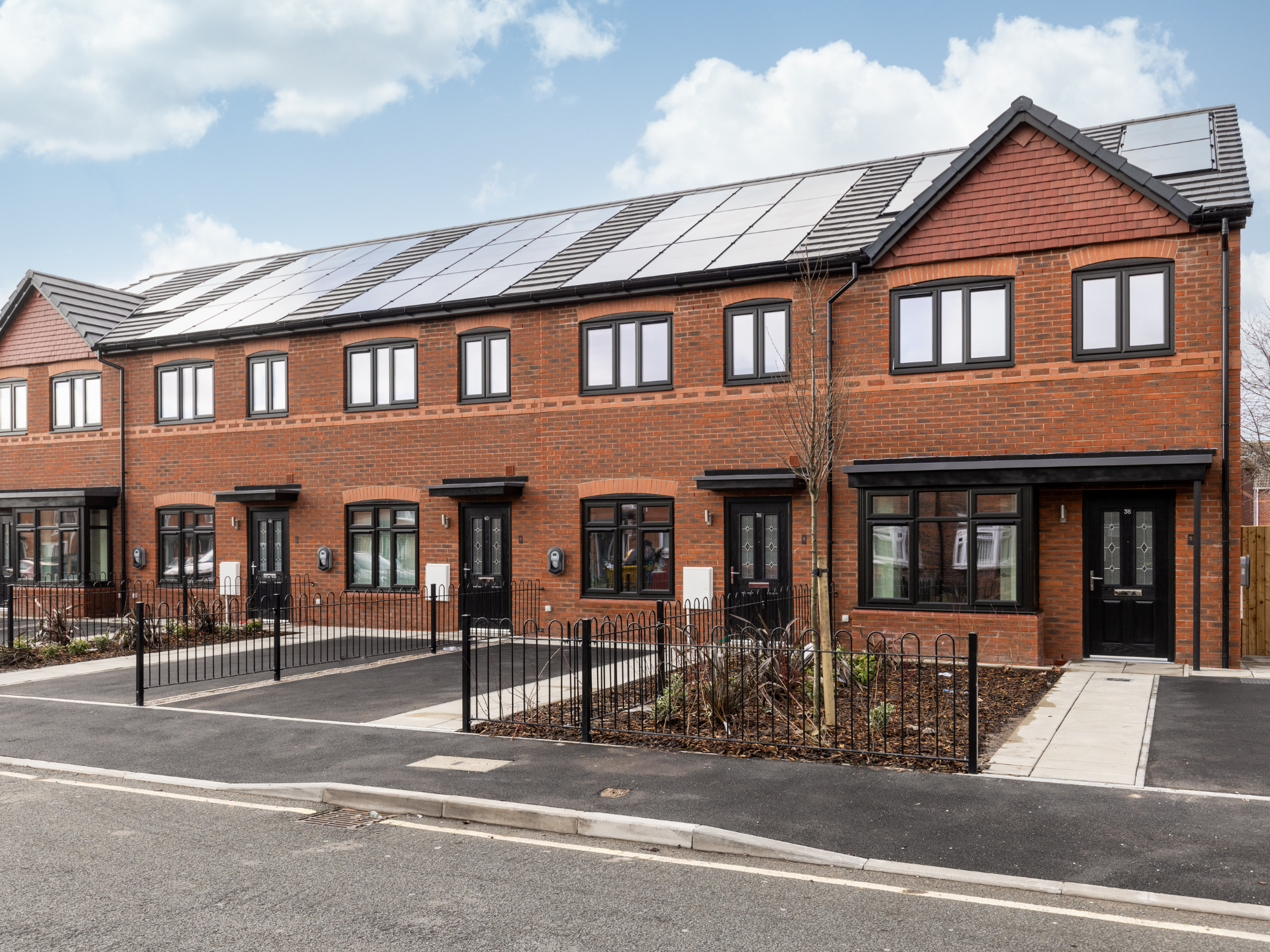 A row of red-brick, new-build homes with driveways in the foreground and a blue sky with white clouds in the background.