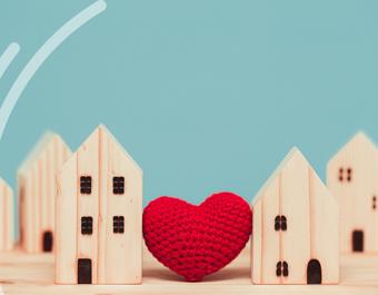 A row of small wooden houses is arranged on a wooden surface against a blue background. A red knitted heart sits between two of the houses in the centre. Three white curved lines appear on the left side.