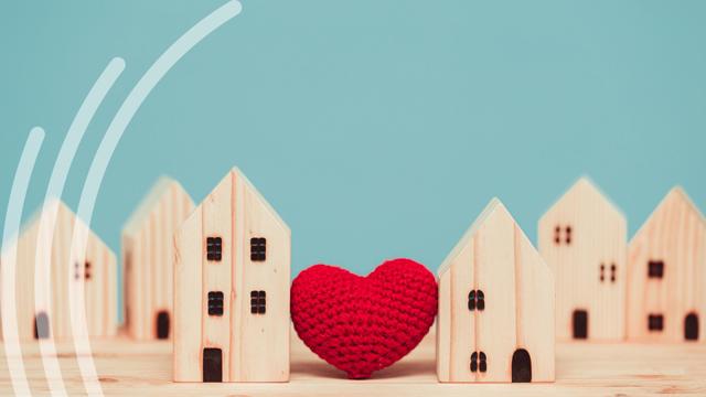 A row of small wooden houses is arranged on a wooden surface against a blue background. A red knitted heart sits between two of the houses in the centre. Three white curved lines appear on the left side.