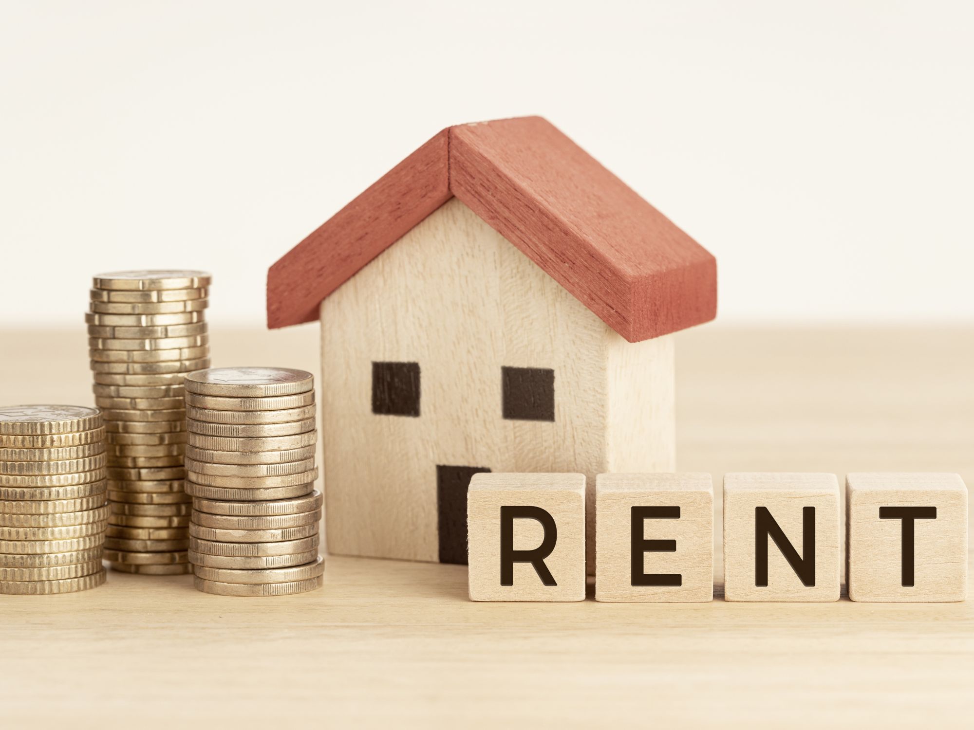 A small wooden house with a red roof sits beside stacks of coins and wooden blocks spelling 'RENT'—symbolizing property rental and rising costs.