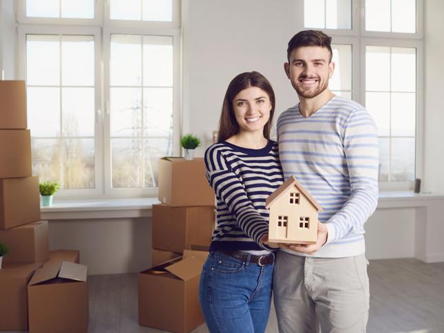 Smiling couple holding a small wooden house model in a bright room with moving boxes, symbolising the transition from renting to buying a home. Smiling couple holding a small wooden house model in a bright room with moving boxes, symbolising the transition from renting to buying a home.