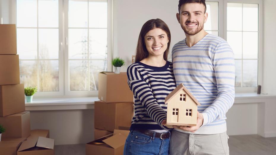 Smiling couple holding a small wooden house model in a bright room with moving boxes, symbolising the transition from renting to buying a home. Smiling couple holding a small wooden house model in a bright room with moving boxes, symbolising the transition from renting to buying a home.