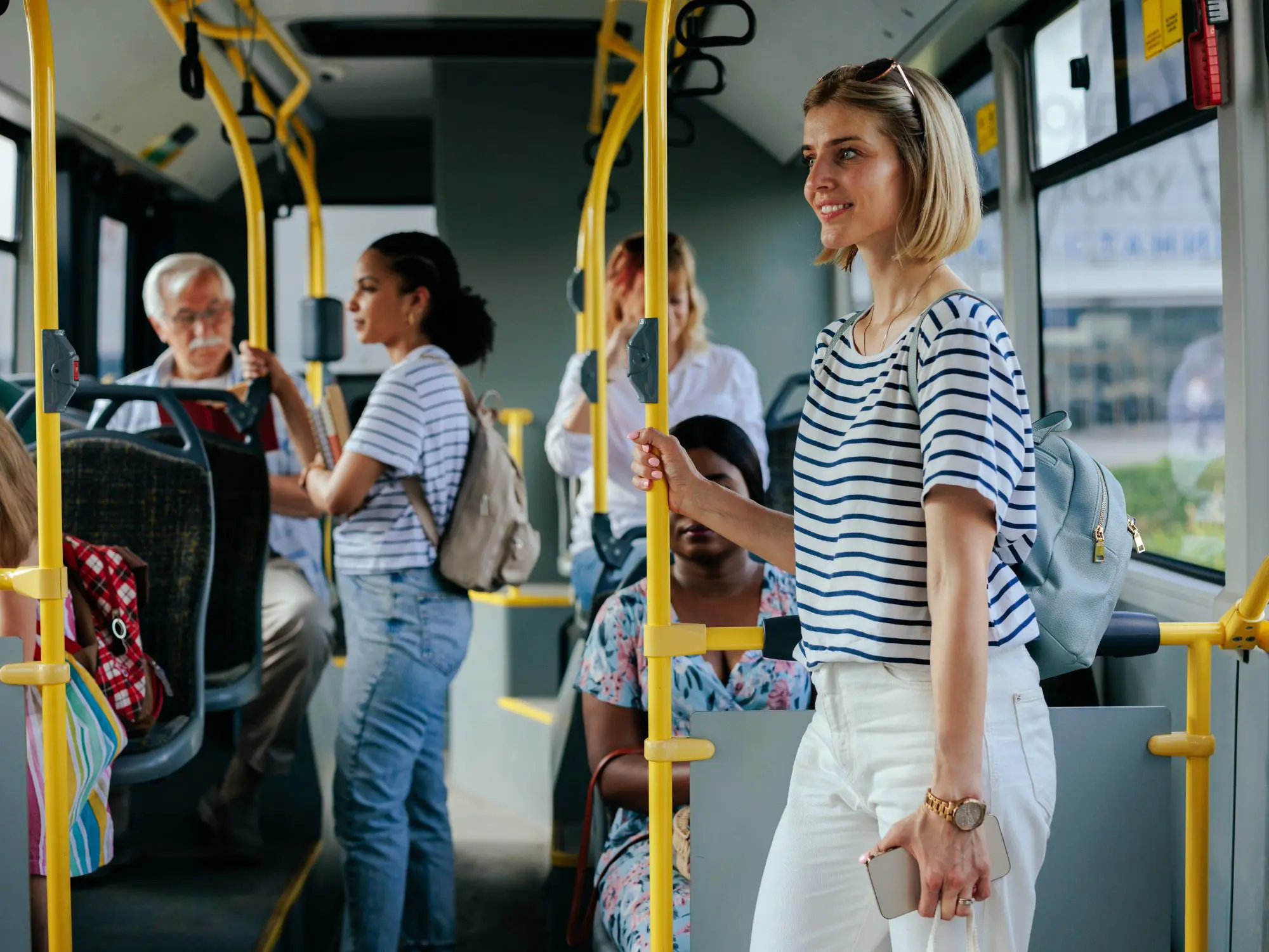 "Interior of a modern bus with passengers standing and sitting, holding onto yellow handrails and engaging in various activities.
