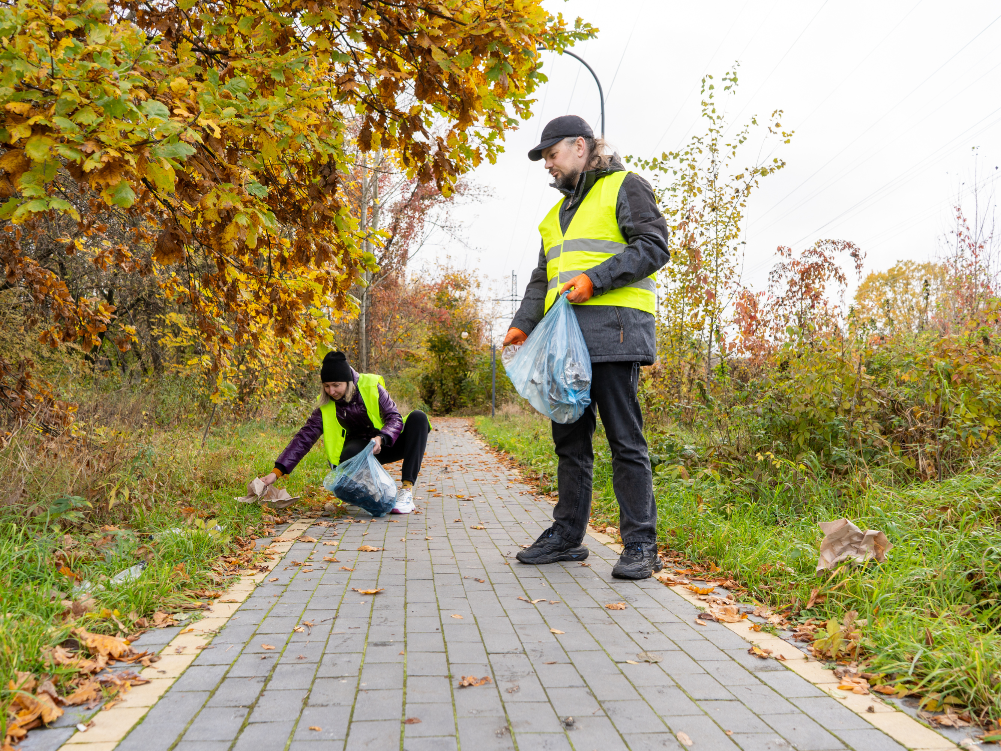 Two people wearing high‑visibility vests are picking up litter along a paved footpath surrounded by autumn trees and overgrown grass. One person is standing and holding a bag of collected rubbish, while the other is crouching near the edge of the path collecting more litter.