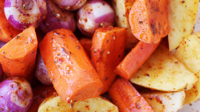 A close-up of chopped vegetables coated in seasoning, including orange carrot pieces, sliced potatoes, and small whole red onions. The vegetables are mixed together and covered in herbs and spices, ready for cooking.