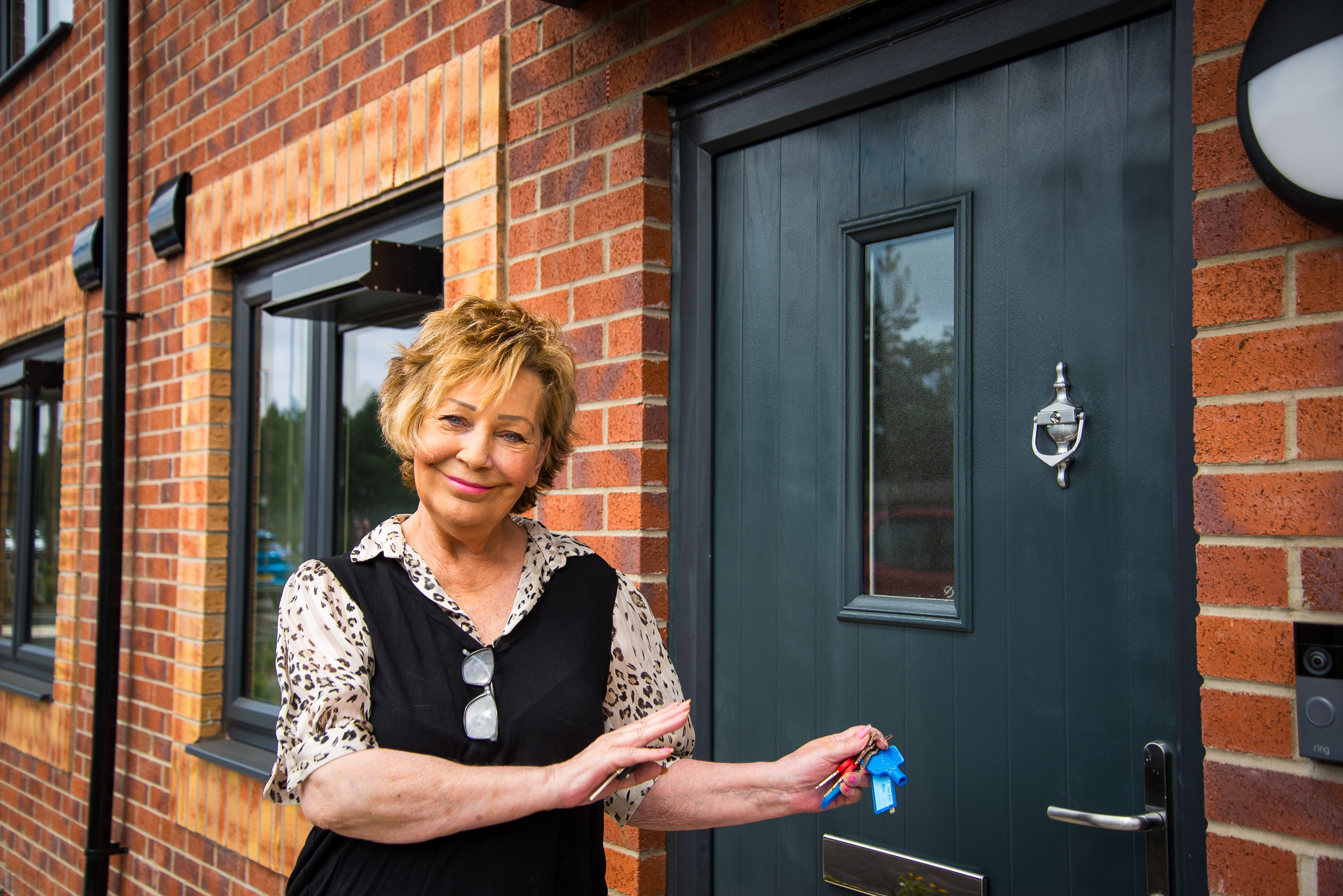A person stands outside a newly built red-brick home, holding a set of keys beside a dark front door. The property features modern black-framed windows, exterior lighting and a door knocker, with the entrance shown in close-up.