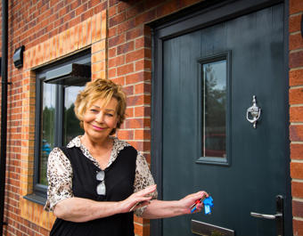 A person stands outside a newly built red-brick home, holding a set of keys beside a dark front door. The property features modern black-framed windows, exterior lighting and a door knocker, with the entrance shown in close-up.