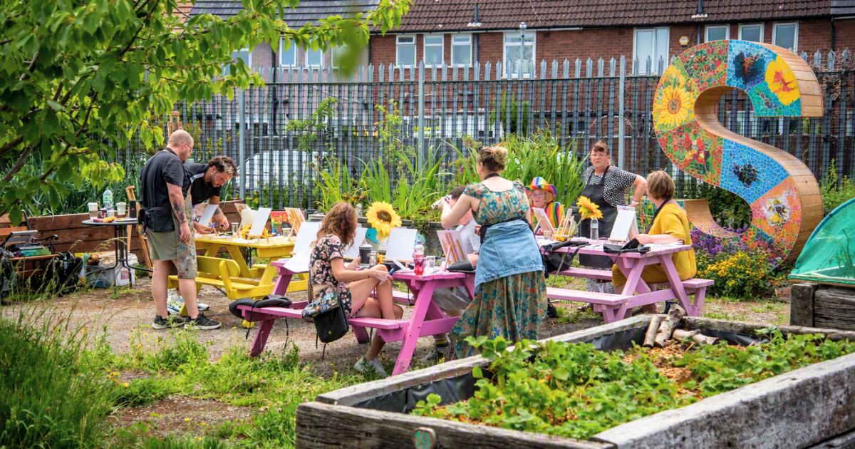 People taking part in an outdoor arts activity around brightly coloured picnic benches in a community garden, beside a large decorative mosaic letter S.