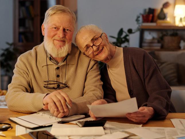 Two older adults sitting at a table with paperwork, a binder, and a calculator, appearing to review documents in a cozy home environment. Two older adults sitting at a table with paperwork, a binder, and a calculator, appearing to review documents in a cozy home environment.