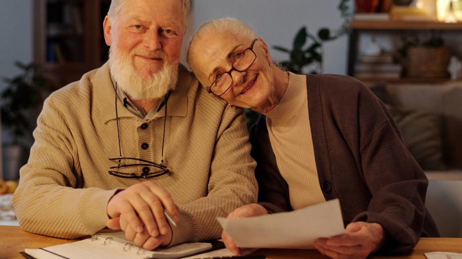 Two older adults sitting at a table with paperwork, a binder, and a calculator, appearing to review documents in a cozy home environment. Two older adults sitting at a table with paperwork, a binder, and a calculator, appearing to review documents in a cozy home environment.