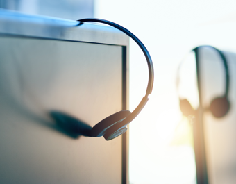 Close-up of a headset hanging on a computer monitor in an office setting, with another similar workstation blurred in the background, representing customer service or call center equipment.