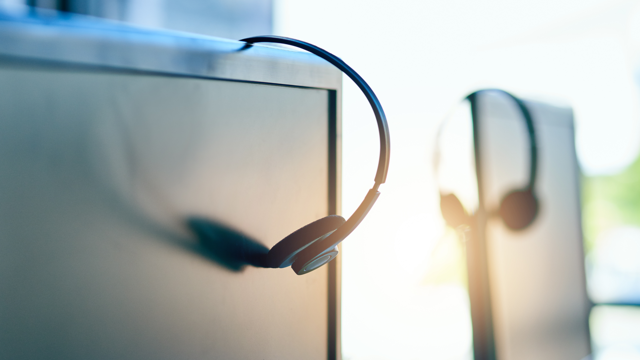 Close-up of a headset hanging on a computer monitor in an office setting, with another similar workstation blurred in the background, representing customer service or call center equipment.