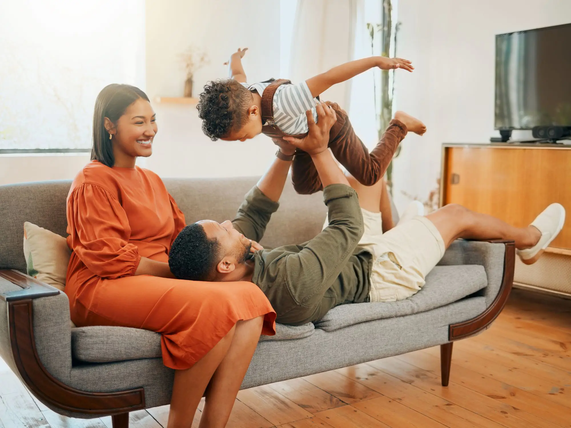 Three people in a living room: one person in an orange dress sits on a couch, another in a green shirt and beige shorts lies on the couch holding up a child dressed in brown overalls and a striped shirt. The room features wooden flooring, a television on a stand, and decorative items in the background.