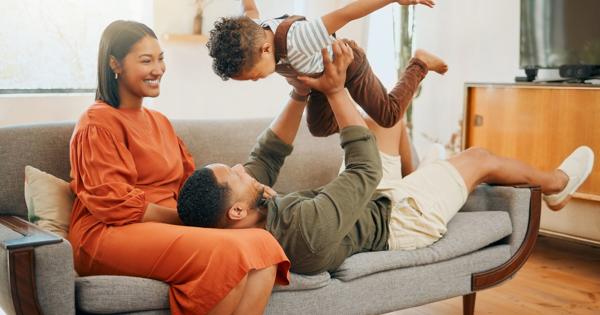 Three people in a living room: one person in an orange dress sits on a couch, another in a green shirt and beige shorts lies on the couch holding up a child dressed in brown overalls and a striped shirt. The room features wooden flooring, a television on a stand, and decorative items in the background.