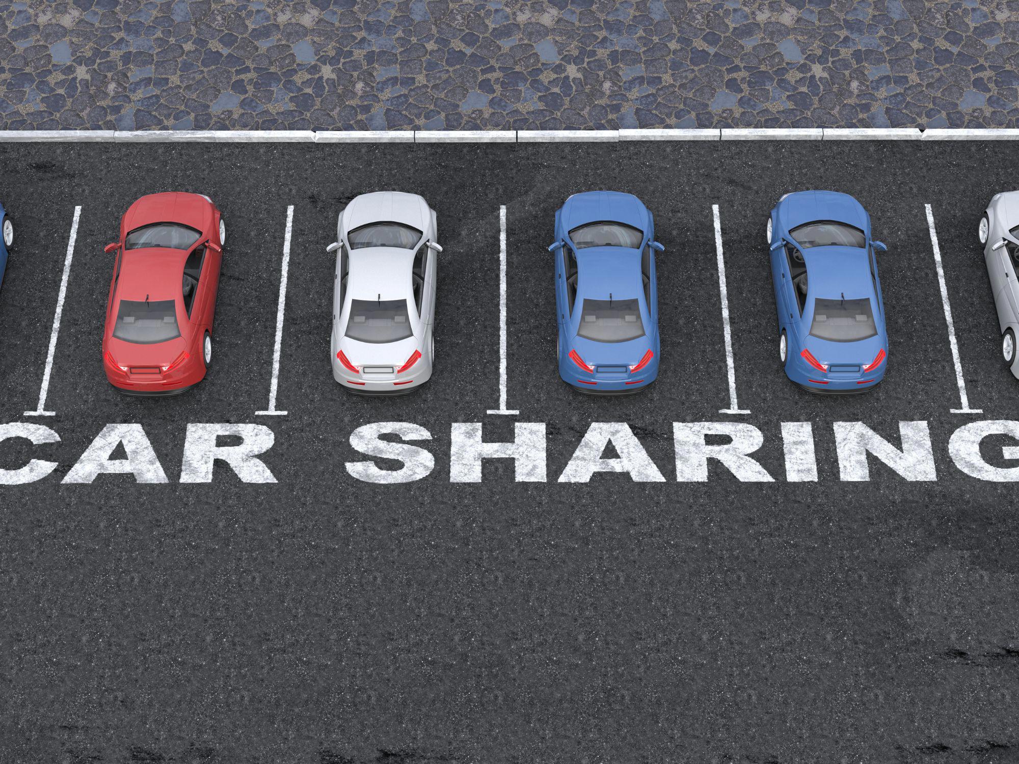 Six cars parked in designated spaces marked CAR SHARING on the asphalt. The vehicles are red, white, and blue, and the setting is a clearly defined parking area.