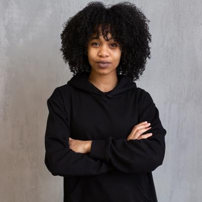 A person with curly hair wearing a black hoodie, standing with arms crossed against a plain gray background.