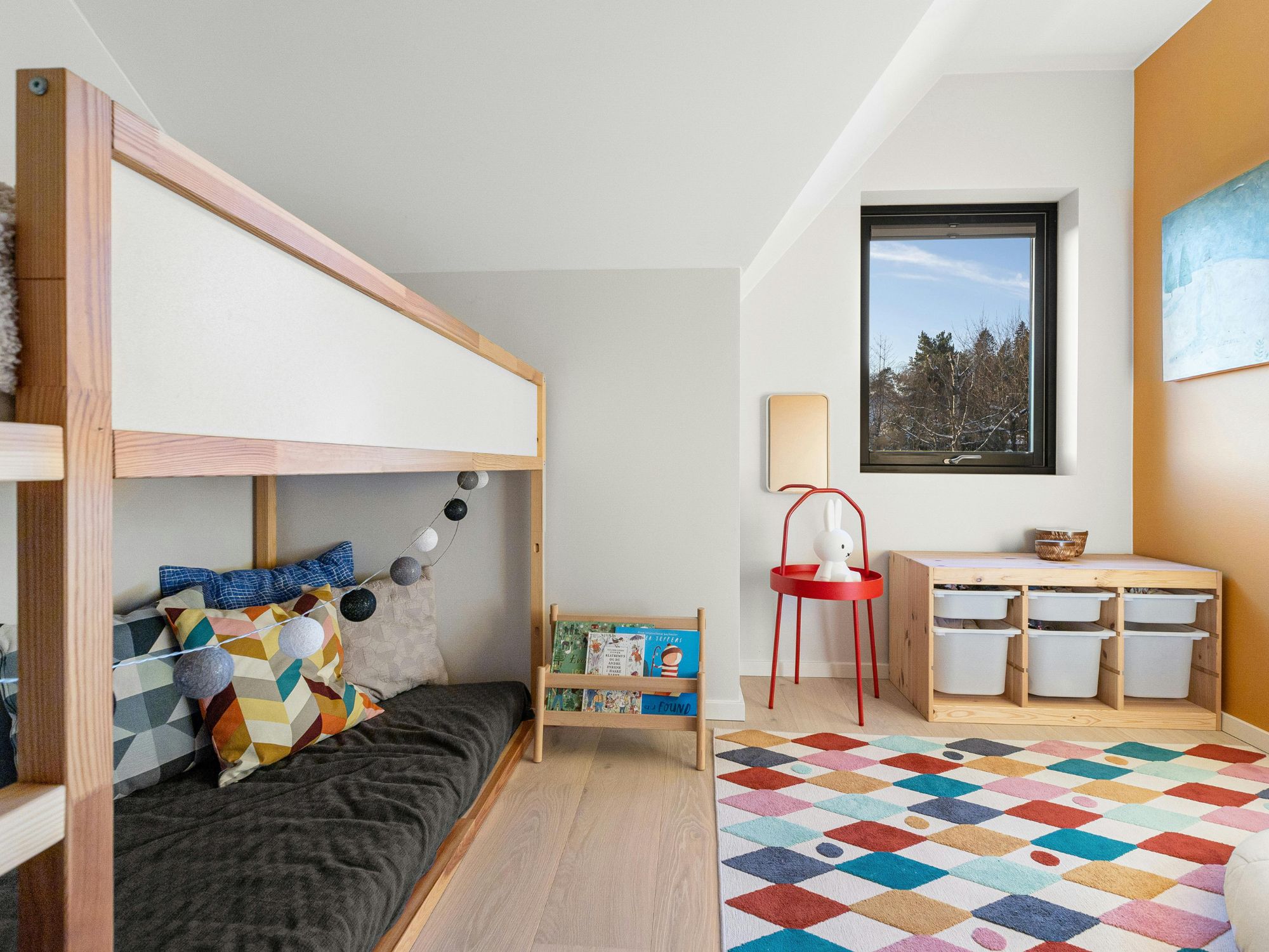 Colorful children's bedroom with a wooden bunk bed, geometric-patterned pillows, string lights, a small bookshelf of children's books, red side table with lamp and stuffed toy, window with storage bins below, and a vibrant diamond-patterned rug.