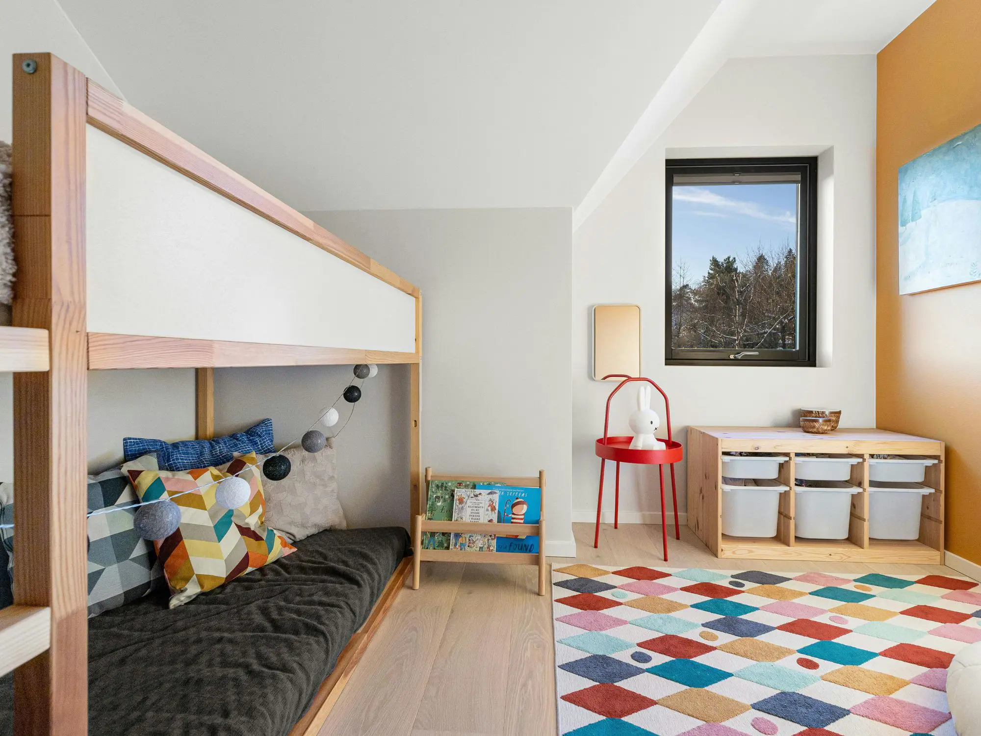 Colorful children's bedroom with a wooden bunk bed, geometric-patterned pillows, string lights, a small bookshelf of children's books, red side table with lamp and stuffed toy, window with storage bins below, and a vibrant diamond-patterned rug.