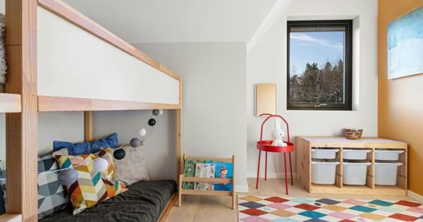 Colorful children's bedroom with a wooden bunk bed, geometric-patterned pillows, string lights, a small bookshelf of children's books, red side table with lamp and stuffed toy, window with storage bins below, and a vibrant diamond-patterned rug.