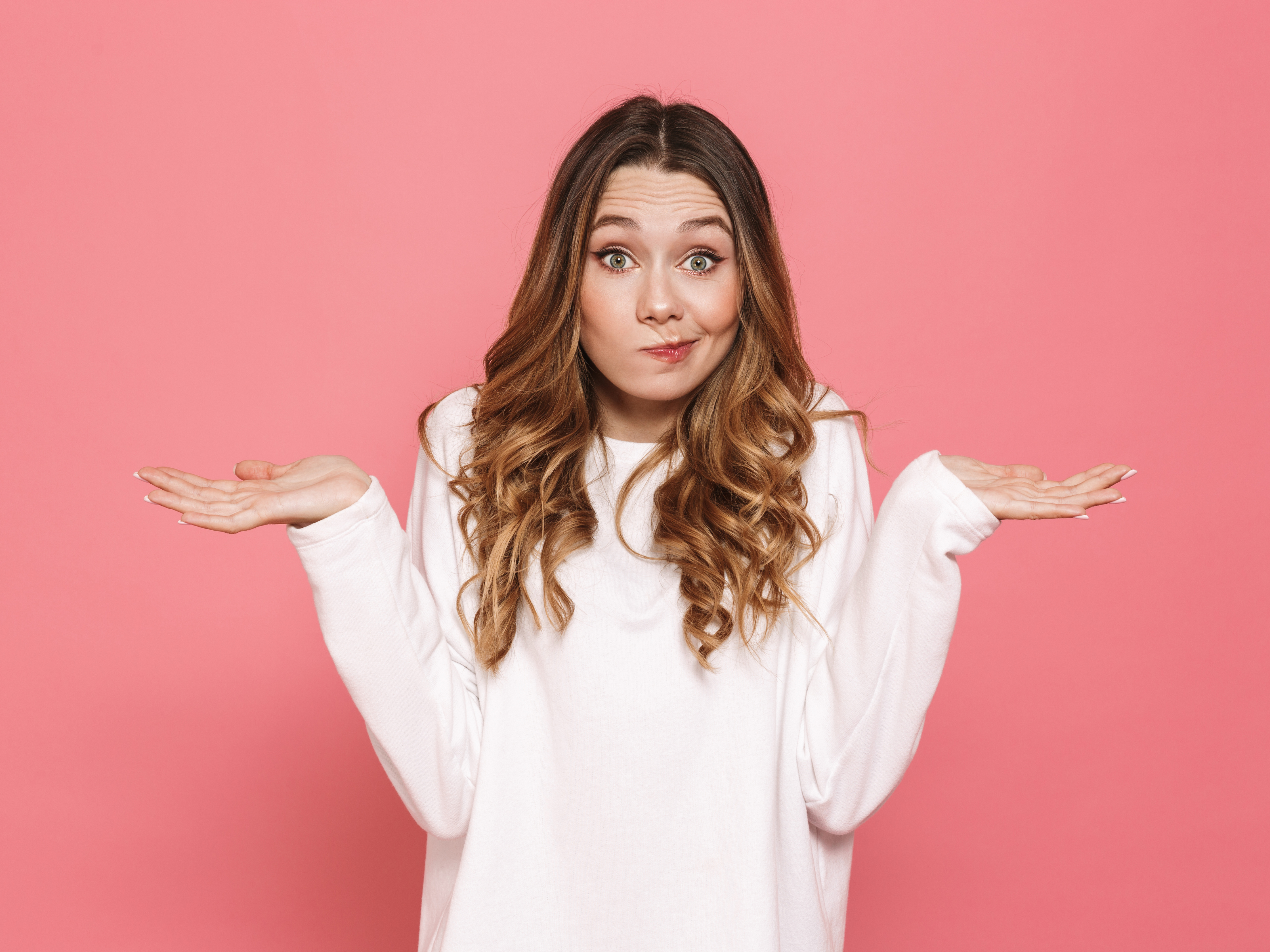 Person in a white top stands against a pink background with both hands raised and palms up in a shrugging pose, looking directly at the camera.